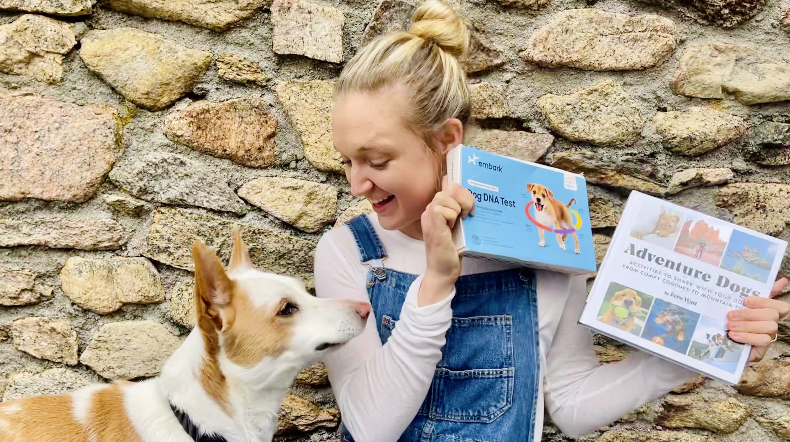 A woman wearing white shirt and overalls looking at her brown and white dog while holding an Embark dog DNA test and a book titled Adventure Dogs.